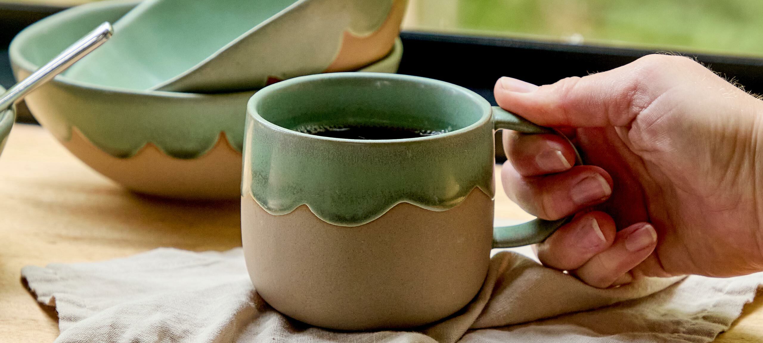 In this image, a green and natural toned mug with black coffee is being picked up by a person's hand