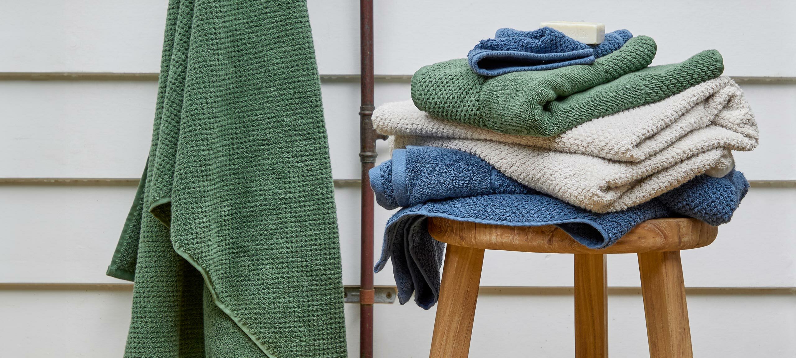 A stack of blue, grey and green towels sit on top of a wooden stool with a green towel hanging in the background. 