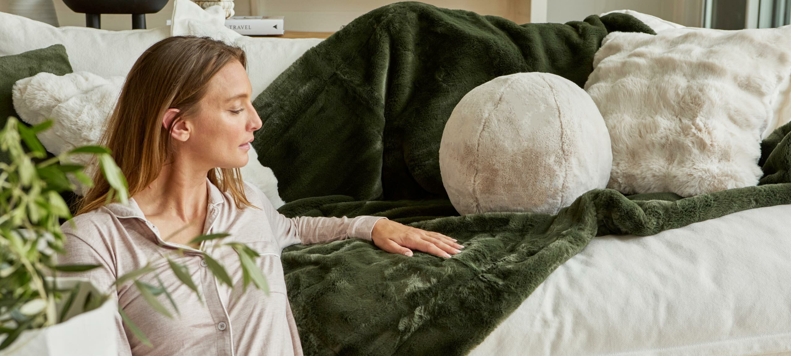 A woman sits in front of a couch. The couch is decorated with a range of natural faux fur cushions and a green faux fur throw. 