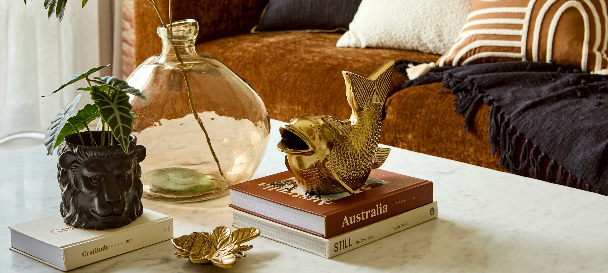 The image shows a marble top coffee table in front of a velvet, tan coloured couch. On the table are a stack of two coffee table books topped with a golden koi statue. Next to the books, a amber vase with a stem and a golden honey bee place sit. On top of a third book sits a black lion head shaped planter with an artificial plant in it. 