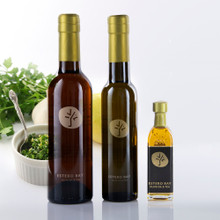 Three sizes of Estero Bay Milanese Gremolata Infused Olive Oil bottles with gold caps, shown with fresh parsley, lemon, garlic, and a bowl of chopped gremolata.