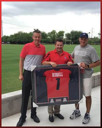 Three men outdoors holding a framed football jersey
