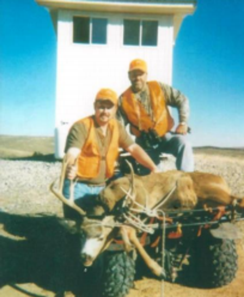 With shooting house in the background 2 hunters and a buck.