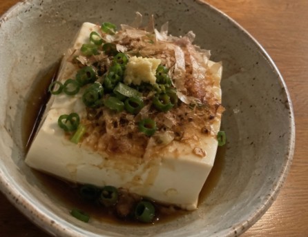 A block of tofu topped with chopped scallions, grated ginger, and bonito flakes, with sauce in a ceramic bowl.