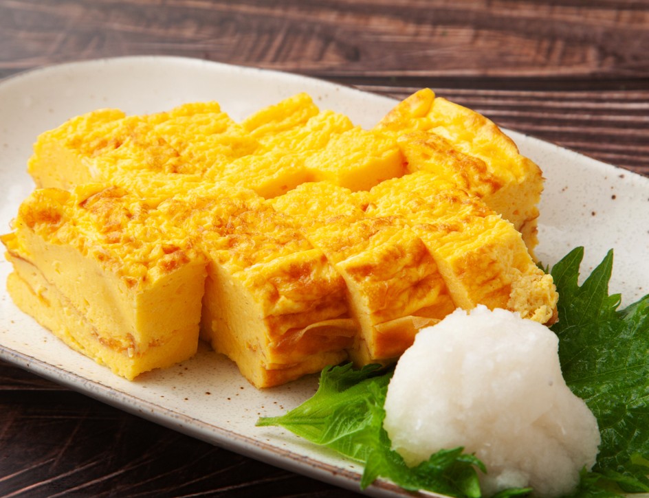 Thick slices of golden Japanese omelet on a cream-colored plate with grated daikon and a green leaf.