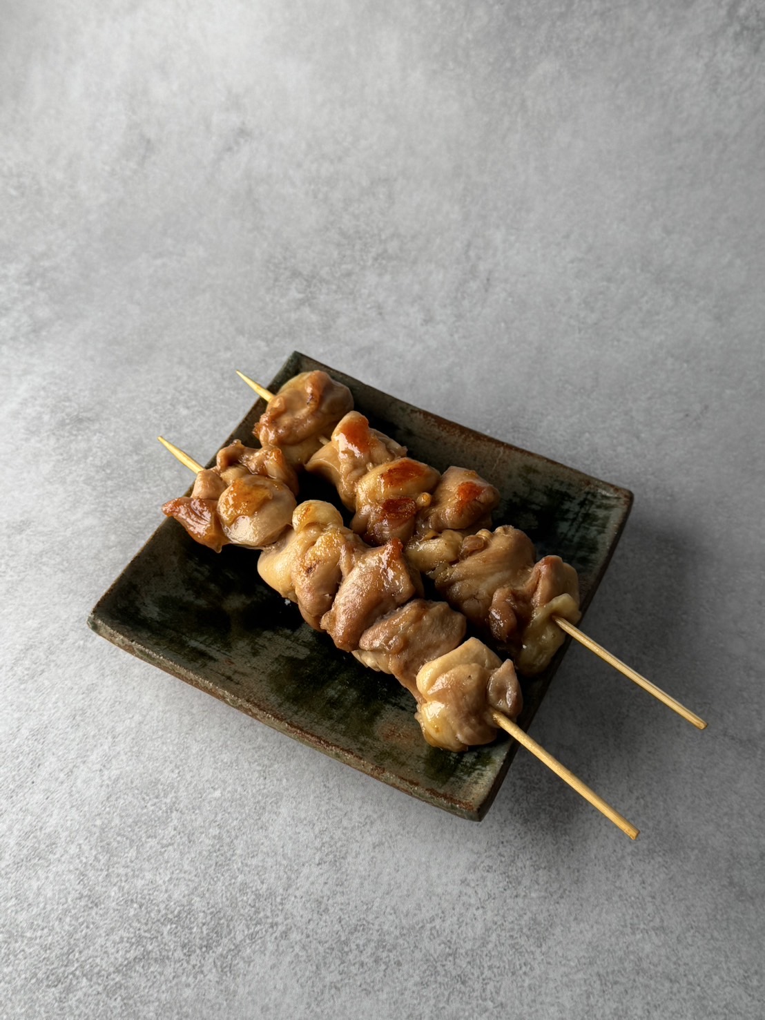 Two grilled chicken skewers on a small rectangular ceramic plate, seen from above at an angle.