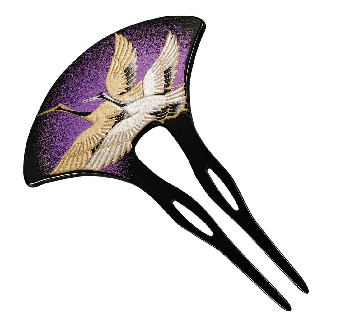 A black lacquered kanzashi hair ornament with two long prongs, its fan-shaped head decorated with two cranes in gold and white against a purple ground