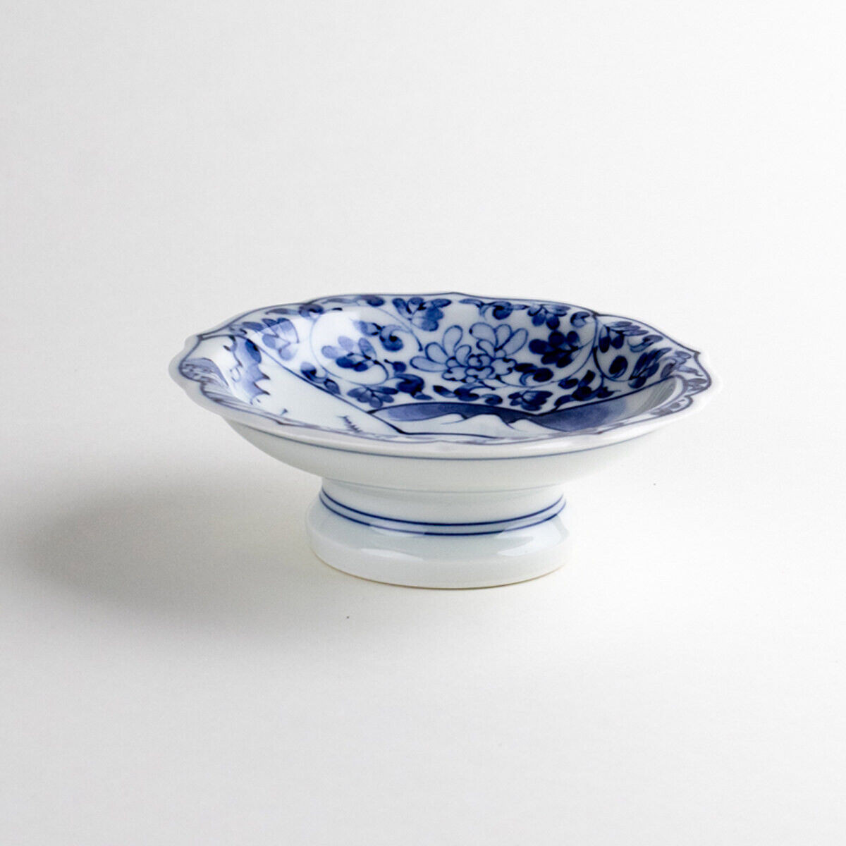A shallow Arita ware small bowl painted in blue and white with a landscape and flowering scrollwork, raised on a small pedestal foot