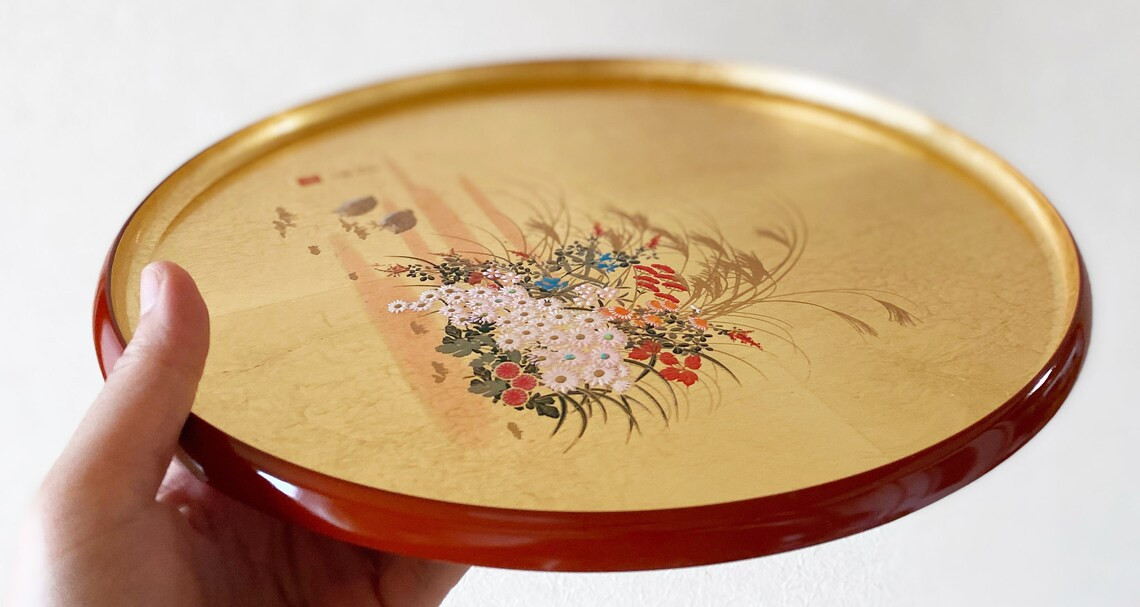 A hand holding a round Yamanaka tray with a gold ground and a painted flower design, tilted toward the camera