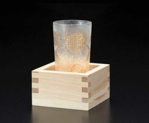 A glass sake cup with gold komon patterning standing beside a cedar masu box
