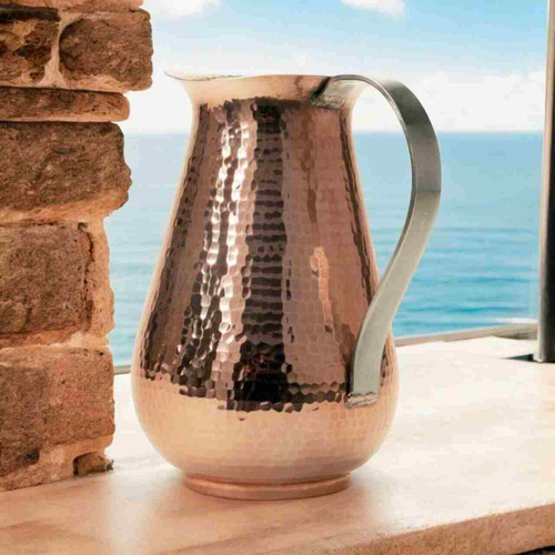 Image shows our elegant Sertodo Bisotun Ayurvedic Copper Water Pitcher, on a stone ledge with an ocean view. Your perfect hydration.