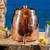Image shows our elegant Sertodo Niagara Ayurvedic Copper Water Dispenser with Lid, on a stone ledge with.an ocean view. Your perfect hydration.