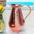 Image shows our elegant Sertodo Bisotun Ayurvedic Copper Water Pitcher, on a stone ledge with.an ocean view. Your perfect hydration.