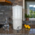 Our AquaCera Terra2 stoneware Gravity Water Filter dispenser on a stone countertop, with a stone wall in the background. To the left, slightly behind the dispenser, is a water carafe with lemons. To the right background is the kitchen window showing a verdant mountain river valley view.