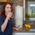 A woman enjoying a refreshing glass of water from our AquaCera Terra2 stoneware Gravity Water Filter dispenser on a stone countertop, with a stone wall in the background. To the right background is the kitchen window showing a verdant mountain view.