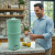 A man enjoying a refreshing glass of water from our AquaCera Terra2 stoneware Gravity Water Filter dispenser on a stone countertop, with a stone wall in the background. To the right background is the kitchen window showing a verdant mountain view.
