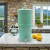 Our AquaCera Terra2 stoneware Gravity Water Filter dispenser on a country cottage kitchen countertop, with a stone wall in the background. To the right background is the kitchen window showing a verdant mountain view.