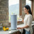 A woman enjoying a relaxing water moment with our AquaCera Terra2 stoneware Gravity Water Filter dispenser on a stone countertop, with a stone wall in the background. To the right background is the kitchen window showing a verdant mountain view.