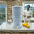 A man enjoying refreshing water from our AquaCera Terra2 stoneware Gravity Water Filter dispenser on a stone countertop, with a stone wall in the background. To the right background is the kitchen window showing a verdant mountain view.