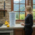 Enjoying a refreshing water moment of reading by our AquaCera Terra2 stoneware Gravity Water Filter dispenser on a stone countertop, with a stone wall in the background. To the right background is the kitchen window showing a verdant mountain view.