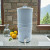 Our AquaCera Terra2 stoneware Gravity Water Filter dispenser on a stone countertop, with a stone wall in the background. To the left, slightly behind the dispenser, is a water carafe with lemons. To the right background is the kitchen window showing a verdant mountain valley view.