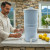 A man enjoying refreshing water from our AquaCera Terra2 stoneware Gravity Water Filter dispenser on a stone countertop, with a stone wall in the background. To the right background is the kitchen window showing a verdant mountain view.