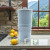 Our AquaCera Terra2 stoneware Gravity Water Filter dispenser on a stone countertop, with a stone wall in the background. To the left, slightly behind the dispenser, is a water carafe with lemons. To the right background is the kitchen window showing a verdant mountain valley view.
