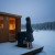 A cedar sauna door slightly open, warm glow spilling out, HUHT cold plunge tub just outside, contrast of heat and cold, steam meeting cold air, balance and recovery.