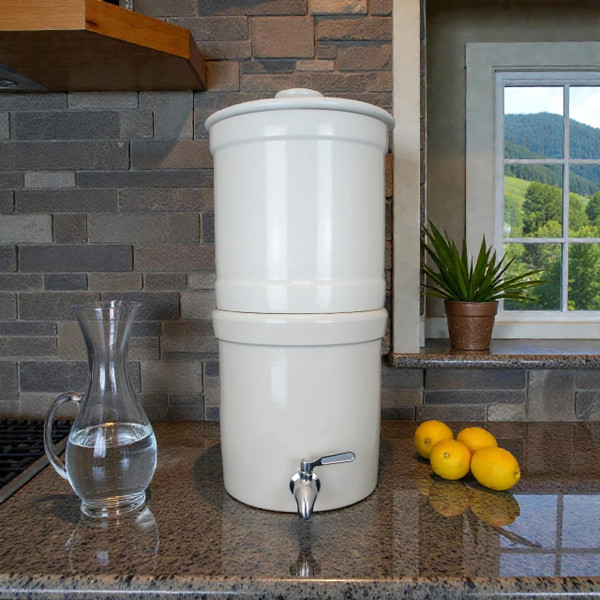 Our AquaCera Terra2 stoneware Gravity Water Filter dispenser on a stone countertop, with a stone wall in the background. To the left, slightly behind the dispenser, is a water carafe with lemons. To the right background is the kitchen window showing a verdant mountain river valley view.