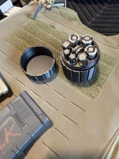 LAPG Tactical Spare Battery Container TDP-RDBATTHOLDER, a round black plastic storage case holding multiple batteries, positioned on a tactical gear surface with a Velcro patch and other equipment around. Thumbnail Image