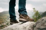 OTB Boots Incline Mid Waterproof Tactical Boot, close-up of rugged brown boots worn on a person's feet while standing on rocky terrain outdoors.