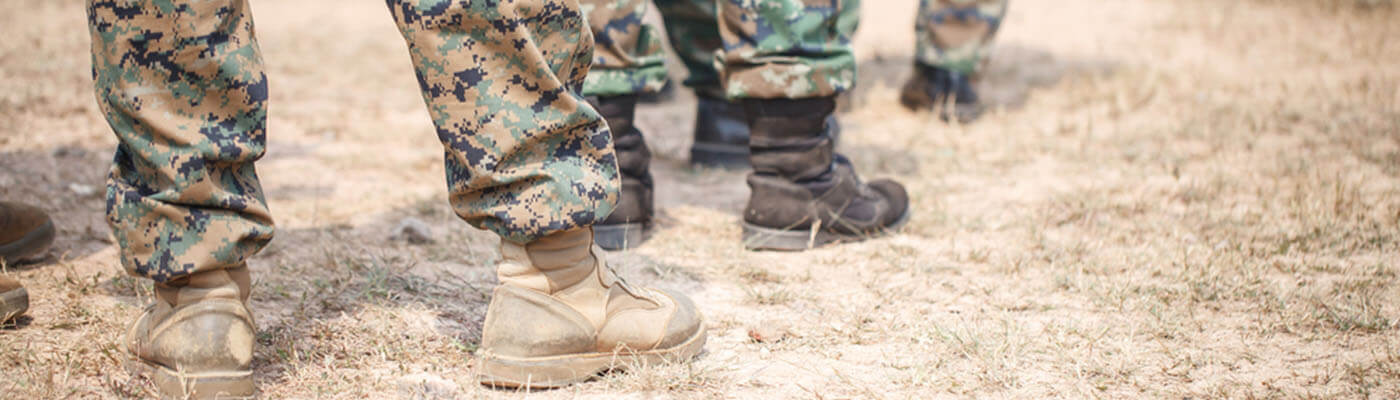 Close-up of soldiers' tactical boots and camouflage pants standing on dry grass in an outdoor setting.