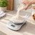 Balanza de cocina digital gris plata con bol transparente donde una mano vierte harina desde un tarro de cristal