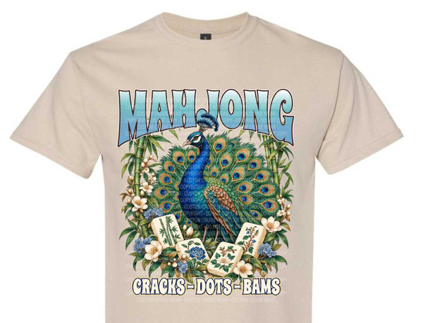 The image is a graphic design printed on a tan t-shirt. The design features a peacock with its tail feathers spread open in a fan shape. The peacock is predominantly blue and green with iridescent accents.

Surrounding the peacock is a lush arrangement of bamboo stalks, white flowers with delicate petals, and small clusters of blue flowers. In the foreground, arranged in a semi-circle around the peacock's feet, are four Mahjong tiles. These tiles display distinct symbols: one with bamboo stalks, one with a blue floral pattern, and two with white floral motifs on a light background.

Above the peacock, in large, stylized blue lettering with a light gradient and a dark outline, is the word "MAHJONG". Below the Mahjong tiles, also in large, stylized lettering with a dark outline and a light yellow-white gradient, are the words "CRACKS - DOTS - BAMS".

The overall color palette is rich and vibrant, with the blues and greens of the peacock and Mahjong tiles contrasting with the natural greens of the bamboo, the whites of the flowers, and the beige of the t-shirt. The composition is centered, with the peacock and the word "MAHJONG" forming the primary focal points. The Mahjong tiles and floral elements add detail and context to the theme. The design has a detailed, illustrative quality. The t-shirt itself is a plain, short-sleeved crew neck.