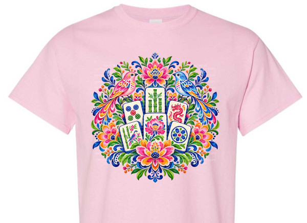 The image displays a pink t-shirt with a vibrant, circular design centered on the chest area. The design features elements inspired by Mahjong tiles and a decorative floral and bird motif.

At the core of the design are several Mahjong tiles arranged in a fan-like pattern. These tiles include: a tile with three bamboo stalks, a tile with a red dragon, a tile with a small bird perched on bamboo, a tile with a blue flower, and a tile with red and green circles. The tiles have a slight tilt and overlap, creating a sense of depth.

Surrounding the Mahjong tiles is an elaborate and colorful arrangement of stylized flora and fauna. Two birds, one pink and one blue, are perched on opposite sides of the design, their elaborate tails fanning out and intertwining with the foliage. The birds have detailed feather patterns. The foliage consists of lush green leaves, vibrant blue swirling elements that resemble peacock feathers or intricate scrolls, and various flowers in shades of pink, orange, yellow, and red. Some smaller red berries are scattered throughout the floral elements.

The overall color palette is rich and saturated, with a strong emphasis on bright, contrasting colors. The background of the t-shirt is a soft, light pink, which makes the vibrant design pop. The composition is circular and symmetrical, with the Mahjong tiles forming a central anchor and the floral elements radiating outwards, creating a balanced and eye-catching arrangement. The style of the artwork appears to be illustrative or painterly, possibly with a watercolor aesthetic, given the soft edges and blended colors in some areas. The t-shirt itself is a standard crew-neck short-sleeved style.