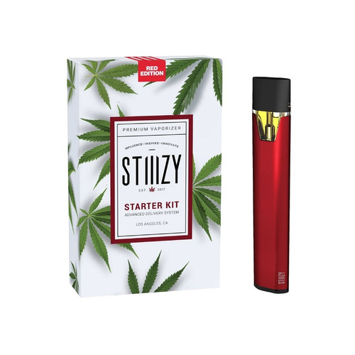 A slim, pen-style STIIIZY Original vape battery in matte black with a gold “V”-shaped airflow window. The battery has a rectangular mouthpiece and clean minimal design—shown beside its branded packaging box featuring stylized palm‑leaf artwork.
