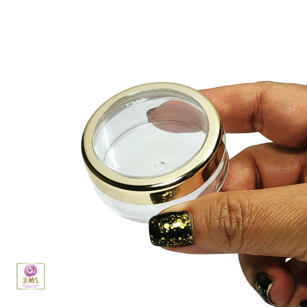 A hand holding  a clear cosmetic jar with gold trim window lid