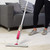 Pink spray mop in use on light wood floor, next to grey sofa.