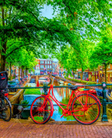  Close up of Red Bike In Amsterdam wooden jigsaw puzzle, featuring a red bicycle parked on a shady brick pathway along a canal.  The canal stretches down through the middle of the image, with buildings and trees running along each side of it. Cars are parked on the side of the road and boats sit floating in the canal, while people walk around Amsterdam. 