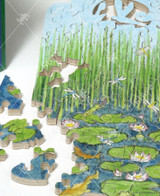  Close up of Every Now And Zen wooden jigsaw puzzle illustrating life on a pond with frogs sitting on lily pads as they try to eat insects, dragonflies buzz through the air, and fish swimming around with tall grass filling the background. 