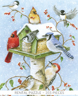  Close-up of Gather In Harmony wooden jigsaw puzzle presenting a green birdhouse feeder mounted on a birch post. Snowflakes fall and cover the top of the feeder. Chickadees, cardinals, a blue jay, a white throated sparrow, and a woodpecker gather to eat the seeds. 