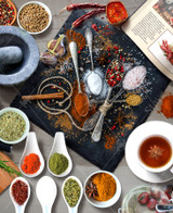  Close up of a stone cutting board covered with a variety of colorful spices, surrounded by small bowls, an open cookbook, and a mortar and pestle. 
