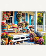  A cozy summer porch scene featuring a white house with blue shutters and a flower-filled porch in full bloom. A bench swing sits in front of a window with a bright quilt and pillows, while a small dog naps in the shade under the steps. 