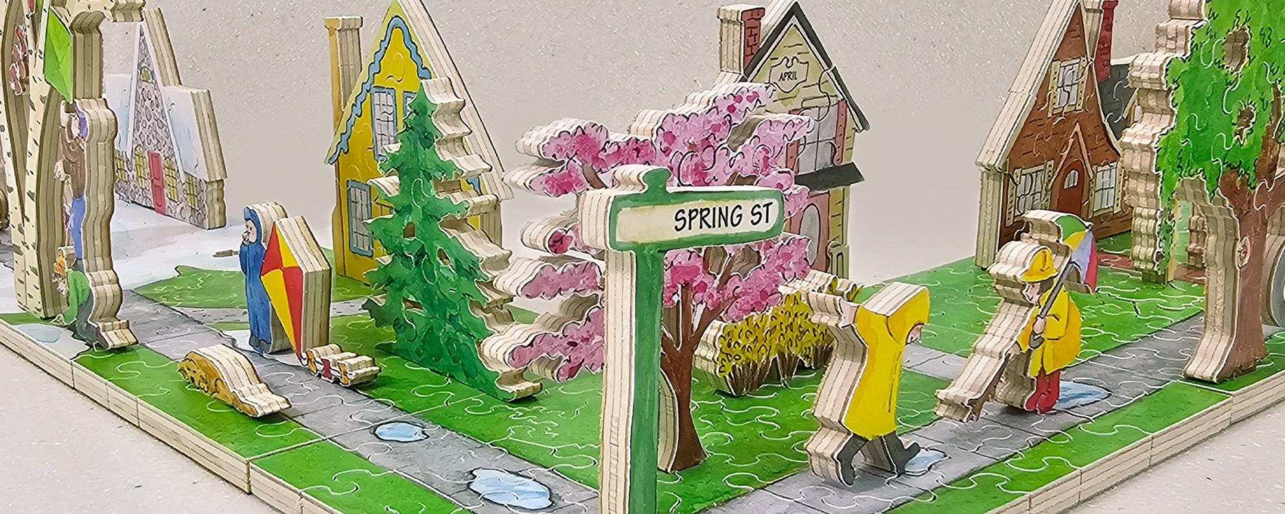 Wooden neighborhood puzzle showing a neighborhood in April. This neighborhood shows people in raincoats, dogs, green and pink trees, and various houses.