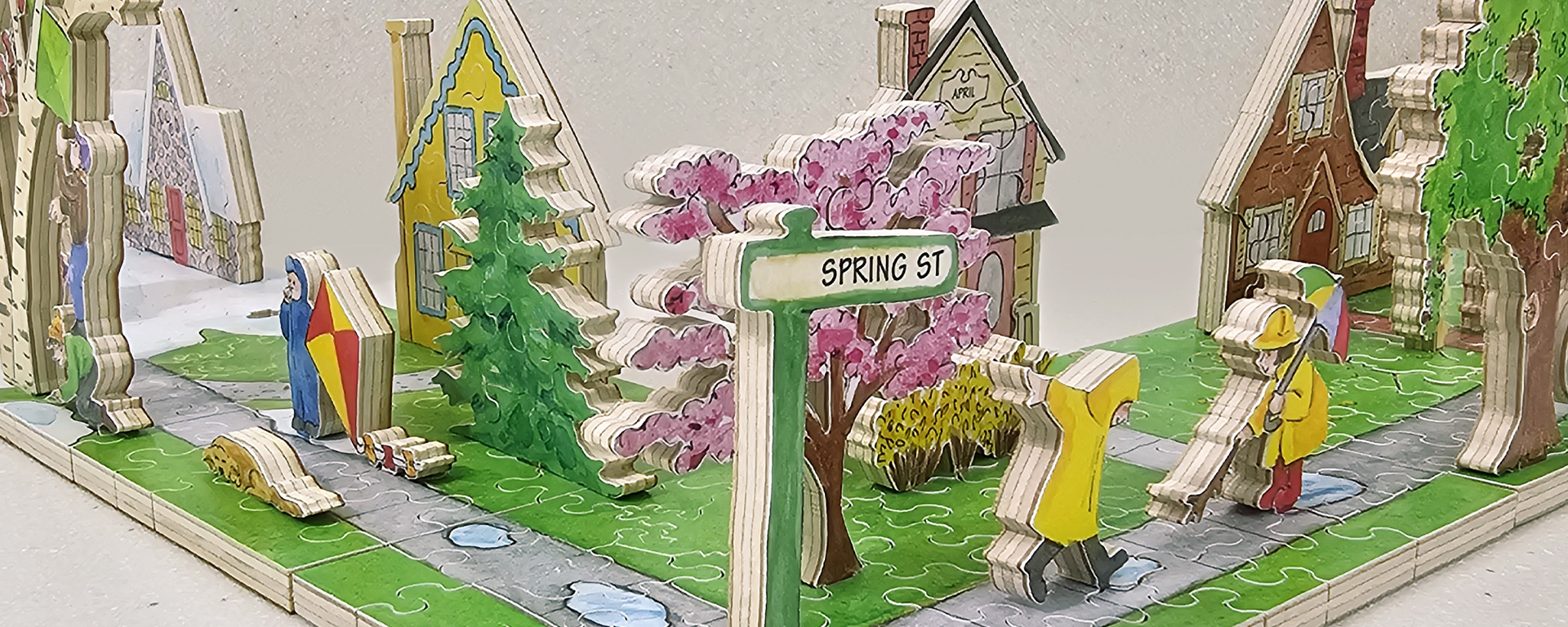 Wooden neighborhood puzzle showing a neighborhood in April. This neighborhood shows people in raincoats, dogs, green and pink trees, and various houses.