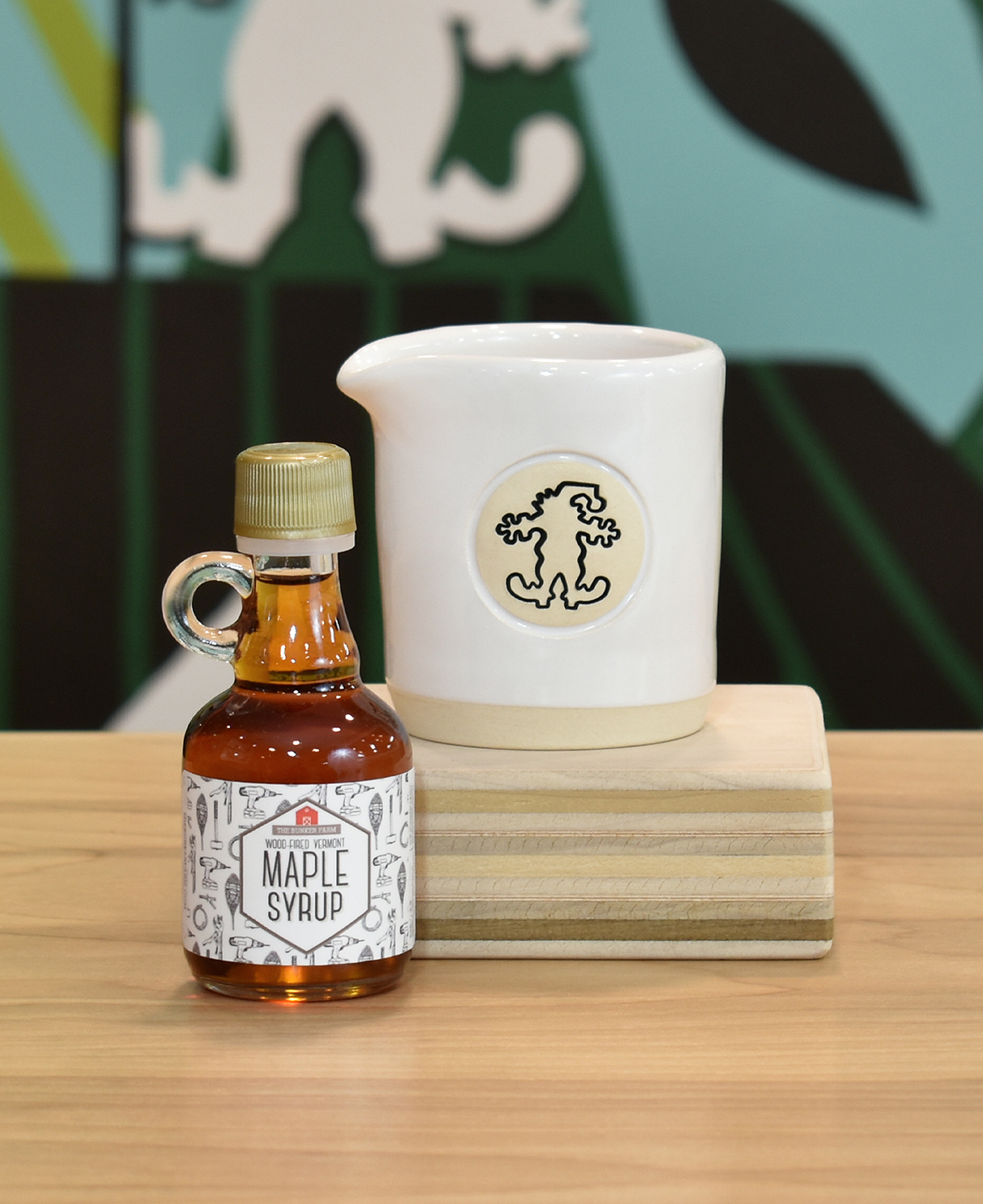Maple syrup pitcher sitting on a wooden block with a bottle of maple syrup on the left side.