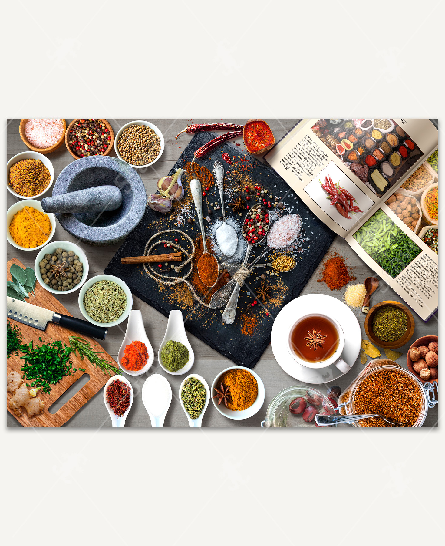 A stone cutting board covered with a variety of colorful spices, surrounded by small bowls, an open cookbook, and a mortar and pestle.