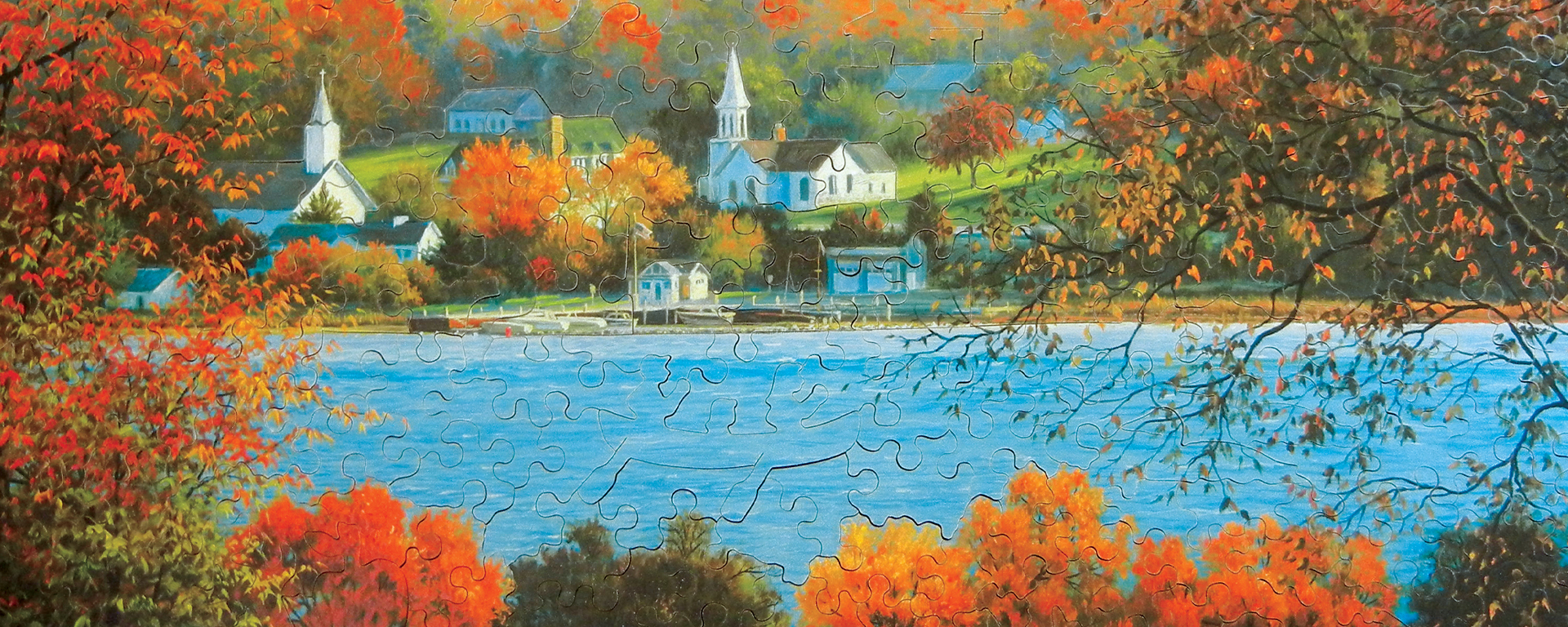 Wooden autumn puzzle of a lake with a quaint town in the background with trees changing their leaves from green to orange.