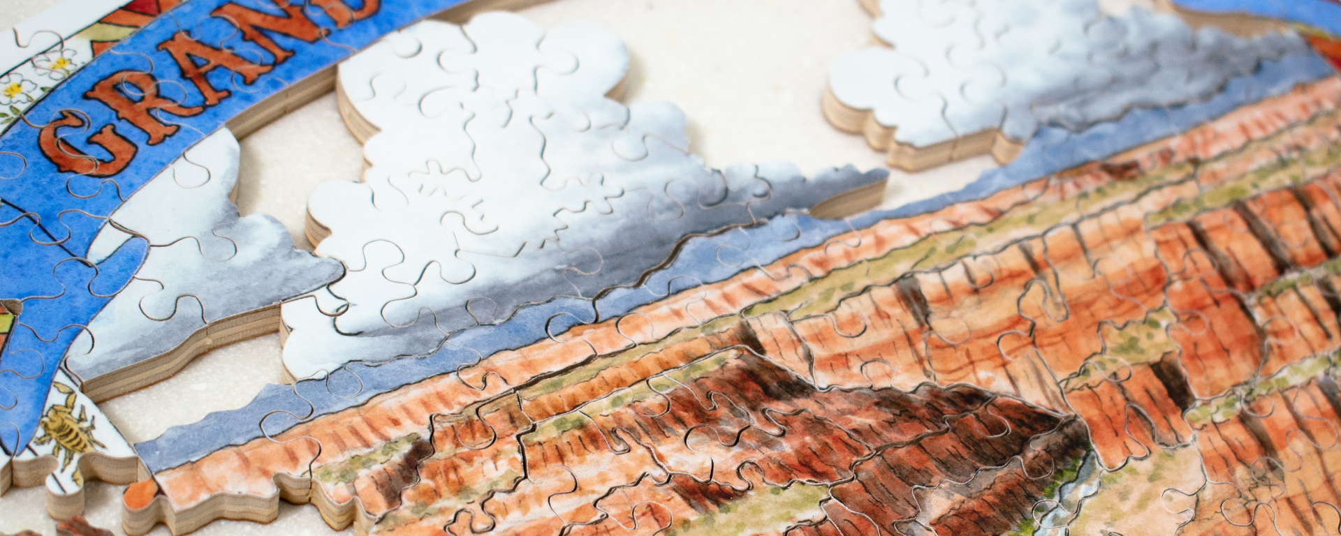 Wooden national park puzzle in progress of the Grand Canyon with clouds in the sky and a blue and orange sign depicting the title of the park with flowers and designs on it.