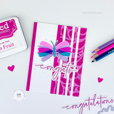 Handmade greeting card with butterfly on it and congrats sentiment on it with crafting materials in background.